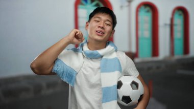 Şehir sokaklarında gülümseyen futbol toplu genç bir adam neşeli bir kutlamada açık havada fular takıyor..