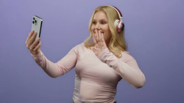 Akıllı telefonlu bir kadın, canlı mor arka plan karşısında kulaklık takarak video kaydediyor, genç enerjisini ve modern teknolojisini göz alıcı, sıradan bir pozda sergiliyor..