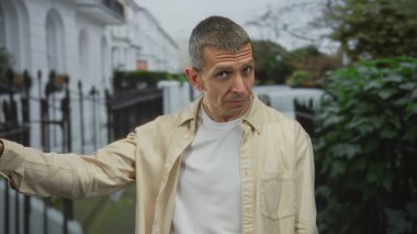 Ciddi yüz ifadeleri olan bir adam, beyaz binaların ve yeşil yeşilliklerin arka planında, serbest bırakılma ya da azarlama önerisi sunan, açık havada jestler yapar..