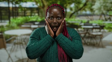 Şehir merkezindeki kafede terasta şaşırmış bir kadın, yeşil kazak giyiyor, şoku, enerjiyi, duyguyu ve şehir ortamını gözler önüne seriyor..