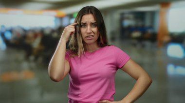 Kadın havaalanında deli gibi jest yapıyor. Kapalı alanda pembe tişört giyiyor. Arka planı bulanık.