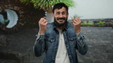 Kot ceketli İspanyol sakallı adam yeşil ağaçlar ve rahat şehir manzarasıyla çevrili bir parkta omuz silkiyor..