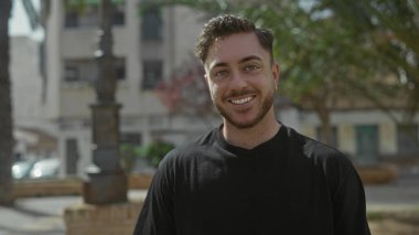 Güneşli bir günde, şehir binaları ve ağaçlarla çevrili, şehir caddesinde duran sakallı genç adam, açık havada hem ciddi hem de gülen yüz ifadeleri sergiliyor..