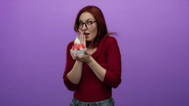 Genç ve kızıl saçlı kadın, yüzünde şaşkınlıkla dondurmayı tutuyor, canlı mor bir duvarın karşısında duruyor, gözlerinde neşe saçıyor, çekici ve anlamlı bir an yaratıyor..