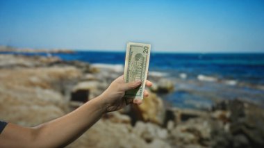 Amerikan dolarlarını deniz kenarında tutan adam, parayı sakin bir kumsal dekoruyla birleştiriyor..