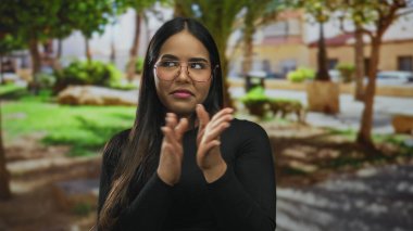 Genç Latin asıllı Latin bir kadının yeşil yeşilliklere karşı poz vermesine rağmen parkta alkışlarken kadın ellerini ayarlıyor..