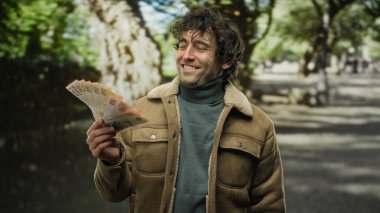 Parkta elinde Türk lirası banknotlarıyla gülümseyen bir adam, açık havada güneşli bir anın tadını çıkararak mutluluğu ve finansal başarıyı yansıtıyor..