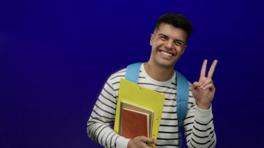 Genç adam gülümsüyor ve mavi arka planda barış işareti olan dosyalar tutuyor, günlük çizgili gömlek ve sırt çantası giyiyor, dostça ve kendinden emin bir görünüm sergiliyor..