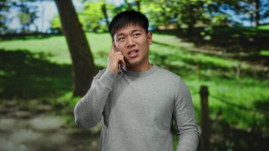 Genç adam güneşli bir parkta telefonla konuşuyor, duygularını canlı bir şekilde ifade ediyor, yemyeşil ağaçlarla çevrili.