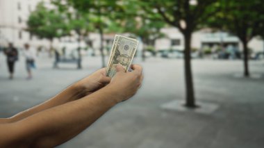 Şehir sokaklarında elinde Amerikan doları olan bir adam ağaçlarla ve bulanık geçmişiyle şehir dışı manzarasını işaret ediyor..