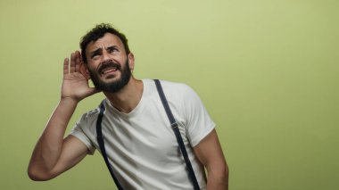 Beyaz gömlek ve pantolon askısı takmış sarı bir fonda dikkatle dinleyen sakallı İspanyol bir adam dikkatli bir ifade ve merak gösteriyor..