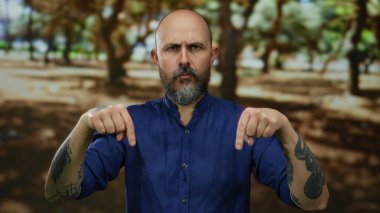 Mavi gömlekli kel bir adam parkta dikiliyor ve kollarında dövmeleri var..