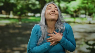 Son sınıf öğrencisi, kır saçlı, elleri göğsünde, doğa ve yeşilliklerle çevrili güneşli bir parkta minnettarlığını ifade eden, dışarıdaki huzur ve mutluluğu somutlaştıran bir kadın..