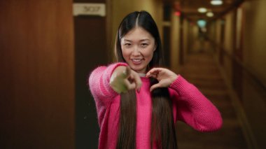 Pembe kazaklı kadın otel koridorunda kalp jesti yapıyor, uzun saçlarıyla gülümsüyor, ev ortamında kendine güveni ve cazibesini sergiliyor, dost canlısı atmosferi vurguluyor..