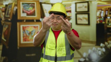 Kasketli ve güvenlik yeleği giyen bir adam duvarları parlak resimlerle dolu bir sanat galerisinde jest yapıyor..
