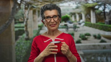 Gri saçlı, kırmızı kazaklı bir kadın yeşil yeşil bir parkta kahve fincanı tutuyor, ağaçlar ve bitkilerle çevrili, dingin ve açık hava manzarası sunuyor..