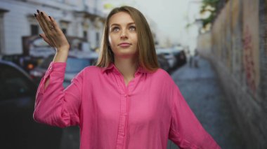 Yoğun bir şehir caddesinde canlı pembe bir tişört giyen bir kadın el hareketleriyle hayal kırıklığı ve kayıtsızlık gösteriyor..