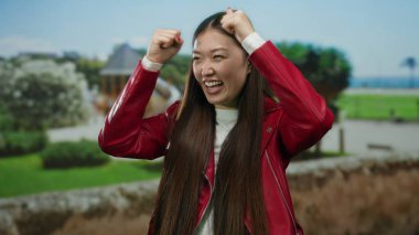 Manzaralı bir parkta zaferi kutlayan heyecanlı bir kadın, kırmızı bir ceket giyiyor, güneşli bir günde yemyeşil bir havayla çevrili mutluluğu ve neşeyi ifade ediyor..