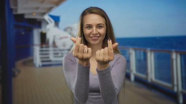 Okyanus manzaralı bir yolcu gemisinde gülümseyen, parmaklarıyla kalp sembolü yapan, uzun kollu bir bluz giyen, barışçıl ve neşeli bir açık hava anı sergileyen bir kadın..