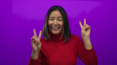 Genç bir kadın gülümsüyor ve barış işaretleriyle poz veriyor, parlak mor bir arka plana karşı, kırmızı bir kazak giyiyor, izole edilmiş bir stüdyoda neşeli bir anı yakalıyor..