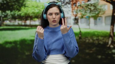 Parkta kulaklık takan bir kadın açık gökyüzünün altında yeşilliklerle çevrili düşünceli bir ifadeyle parmaklarını sayıyor..