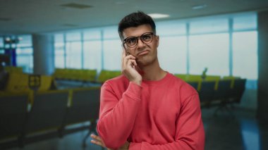Havaalanı terminalinde telefonla konuşan genç adam düşünceli ve düşünceli görünüyor. Gözlük takıyor ve arka planda gri koltuklu pembe süveter giyiyor. Hafif doğal ışık altında.