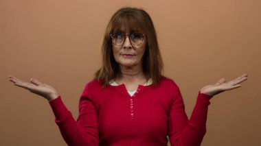 Kırmızı kazaklı yaşlı İspanyol kadın, kollarını kahverengi izole bir duvar arkasına uzatarak düşünceli bir şekilde poz veriyor, tefekkür ve karar verme sürecini anlatıyor..