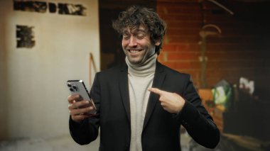 Kapalı alanda gülümseyen yakışıklı bir adam inşaat alanında cep telefonunu işaret ederek şık kıyafetler giyerek ve heyecanını belli eden loş kentsel ortama güvenerek gösteriyor..
