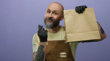 Sakallı, eldiven ve önlük takan kel bir adam, elinde kese kağıdıyla, izole edilmiş mor arka planda rahatça poz veriyor..