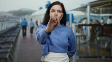 Esmer bir kadın yolcu gemisinin güvertesinde, arka planda bir sürü insan ve şezlongla el kol hareketi yapıyor.