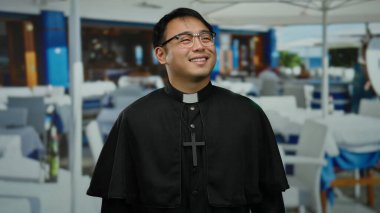 Açık hava restoranında masaların arka planında duran gülümseyen bir rahip ve şehir ortamını gözler önüne seren insanlar.