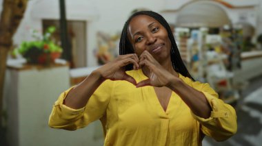 Kıvırcık saçlarıyla gülümseyen ve kalbi dışarıda, şehir mutluluğunu gösteren sarı bluz giyen bir kadın.