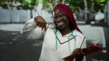 Beyaz tişörtlü bir kadın şehir sokaklarında elinde bir pano, gözlük ve steteskop tutarken sevinçle gülümserken, şehir ortamında profesyonel sağlığa işaret ediyor..