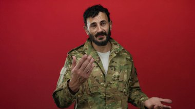 Kırmızı arka planda askeri üniformalı İspanyol sakallı bir adam gururlu ve otoriter bir şekilde kameraya bakıyor.