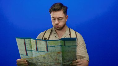 Sırt çantası ve açık havada modern seyahat deneyimini tasvir eden günlük kıyafetlerle gezerken, elinde sakal tutan genç adam mavi arka plan karşısında şaşkın görünüyor..