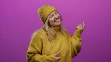 Genç sarışın kadın hardal süveter giyip pembe arka planda gitar taklidi yapıyor..