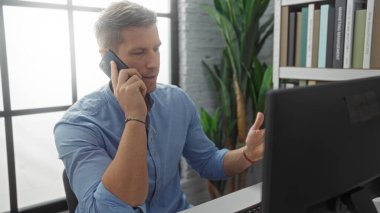 Ofis masasında telefonla konuşan genç bir adam, modern işyerinde bilgisayarla konuşuyor. Etrafı kitaplar ve yeşilliklerle çevrili. Kapalı alanda profesyonel bir atmosfer sergiliyor..