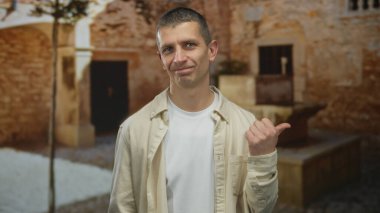 Sıradan kıyafetler giyen bir adam taştan bir arka planı olan bir sokakta dikiliyor, gülümsüyor ve işaret ediyor, güneşli bir günde rahat bir hava veriyor..