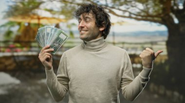 Parkta elinde bangladeş banknotları olan İspanyol bir adam başparmağıyla gülümsüyor ve işaret ediyor. Arka planda ağaçlar, açık havada güneşli bir gün..