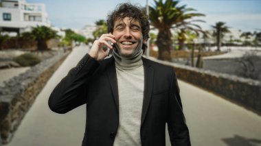 Güneşli bir şehir sokağında palmiye ağaçlarıyla konuşurken gülümseyen genç adam, siyah bir ceket ve balıkçı yaka giyiyor, mutluluğu ve modern şehir yaşam tarzını somutlaştırıyor..