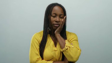 Kadın, kollarını düz beyaz bir arka plana katlamış, parlak sarı bir gömlek giymiş, Afrika kökenli ve sakin tavırlar sergiliyor..