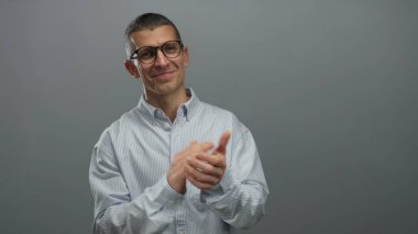 Gözlüklü ve çizgili tişörtlü bir adam izole edilmiş gri duvar arkaplanına karşı alkış tutuyor. Onayını ve pozitifliğini çağdaş bir ortamda, kendine güvenen bir gülümsemeyle ifade ediyor.
