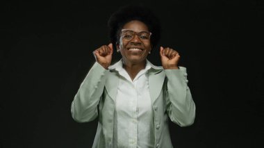 Gözlüklü ve yeşil takım elbiseli bir kadın, kendinden emin bir şekilde gülümsüyor. Olumlu ve neşeyi temsil ediyor..