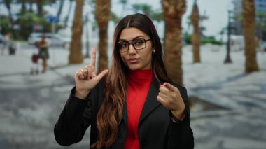 Gözlüklü İspanyol kadın, palmiye ağaçlarıyla kaplı güneşli bir caddede kamerayı işaret ederken 