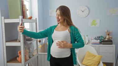 Bebek odasında oyuncak ve beşikle selfie çeken hamile bir kadın. Evdeki mutlu anları yakalıyor. Heyecan ve annelik beklentilerini dile getiriyor.