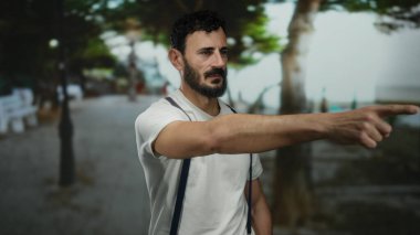 Pantolon askısı takan İspanyol bir adam önde duruyor, arka planda ağaçlar ve banklar olan bir şehir caddesinde duruyor..