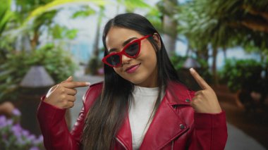 Kırmızı ceketli, güneş gözlüklü bir kadın. Parkta gülümsüyor. Güneş gözlüklerini iki eliyle vurguluyor..