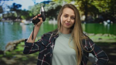 Parktaki genç bir kadın, yeşil açık havada, doğal çevreyi ve zanaatkarlık kontrastını gözler önüne seren sakin bir ifadeyle matkabı tutuyor..