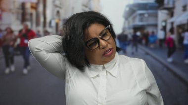Şehir caddesinde beyaz gömlek giyen Latin bir kadın, boyun ağrısı var gibi görünüyor, kentsel arka plan birden fazla yayanın hareket ettiğini gösteriyor..