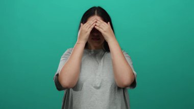 Gözlüklü ve uzun saçlı bir kadın, parlak yeşil bir arka plana karşı şaşkınlığını dile getiriyor, gündelik tarzda giyiniyor ve çevreden soyutlanmış, kıvrımlı figürler sergiliyor..